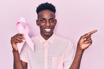 Young african american man holding pink cancer ribbon smiling happy pointing with hand and finger...