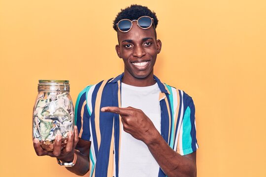Young african american man holding jar with savings smiling happy pointing with hand and finger