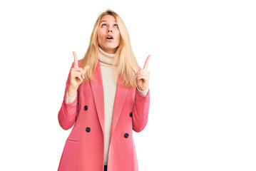 Young beautiful blonde woman wearing elegant clothes amazed and surprised looking up and pointing with fingers and raised arms.