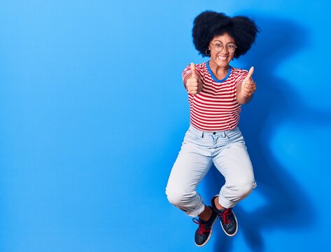 Young beautiful african american woman wearing casual clothes and glasses smiling happy. Jumping with smile on face doing ok sign with thumbs up over isolated blue background