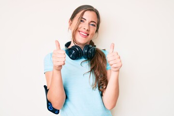 Beautiful young caucasian woman wearing gym clothes and using headphones approving doing positive gesture with hand, thumbs up smiling and happy for success. winner gesture.