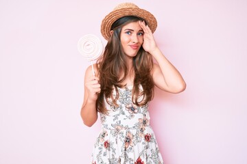 Beautiful young caucasian woman wearing summer hat and lollipop stressed and frustrated with hand on head, surprised and angry face
