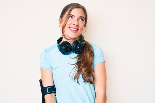 Beautiful young caucasian woman wearing gym clothes and using headphones looking away to side with smile on face, natural expression. laughing confident.