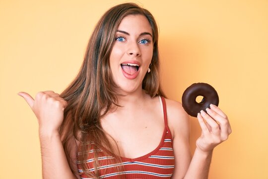 Beautiful young caucasian woman holding donut pointing thumb up to the side smiling happy with open mouth