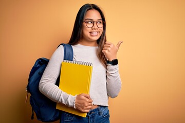 Young smart asian student girl wearing backpack holding notebook over yellow background pointing and showing with thumb up to the side with happy face smiling