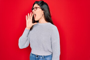 Young beautiful brunette woman wearing casual sweater over red background shouting and screaming loud to side with hand on mouth. Communication concept.