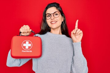 Young beautiful girl holding first aid kit medical box over red background surprised with an idea...