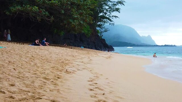 HD Hawaii Kauai slow motion pan left to right of few people on beach to person in distance sitting in ocean waves with Puff the Magic Dragon of Hanalei (Honah Lee) mountainous coastline in background
