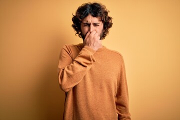 Fototapeta na wymiar Young handsome man with beard wearing casual sweater standing over yellow background smelling something stinky and disgusting, intolerable smell, holding breath with fingers on nose. Bad smell