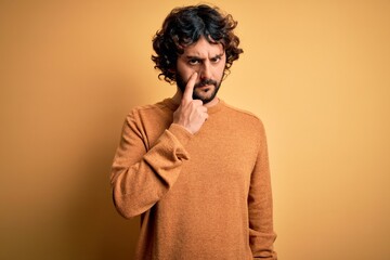 Fototapeta na wymiar Young handsome man with beard wearing casual sweater standing over yellow background Pointing to the eye watching you gesture, suspicious expression
