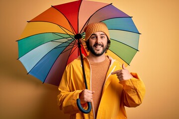 Young handsome man with beard wearing raincoat for rainy day holding colorful umbrella with...