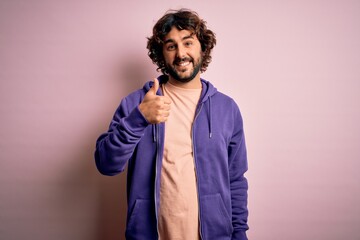 Young handsome sporty man with beard wearing casual sweatshirt over pink background doing happy thumbs up gesture with hand. Approving expression looking at the camera showing success.