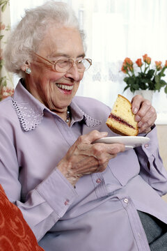 An old bespectacled woman sitting on the couch eating cake
