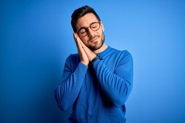 Young handsome man with beard wearing casual sweater and glasses over blue background sleeping tired dreaming and posing with hands together while smiling with closed eyes.