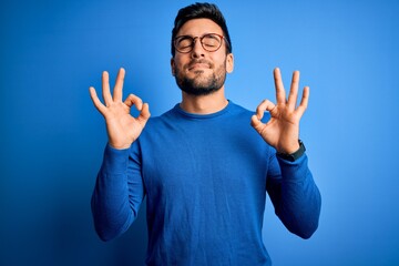 Naklejka premium Young handsome man with beard wearing casual sweater and glasses over blue background relaxed and smiling with eyes closed doing meditation gesture with fingers. Yoga concept.