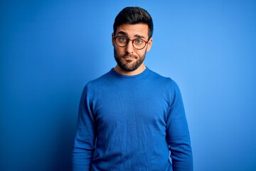 Young handsome man with beard wearing casual sweater and glasses over blue background skeptic and...