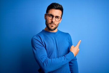 Young handsome man with beard wearing casual sweater and glasses over blue background Pointing with hand finger to the side showing advertisement, serious and calm face