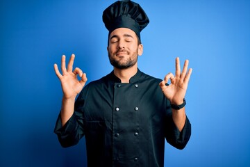 Young handsome chef man with beard wearing cooker uniform and hat over blue background relax and smiling with eyes closed doing meditation gesture with fingers. Yoga concept.