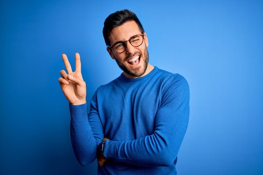 Young handsome man with beard wearing casual sweater and glasses over blue background smiling with happy face winking at the camera doing victory sign with fingers. Number two.