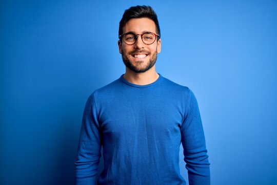 Young handsome man with beard wearing casual sweater and glasses over blue background with a happy and cool smile on face. Lucky person.