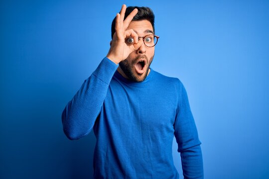 Young handsome man with beard wearing casual sweater and glasses over blue background doing ok gesture shocked with surprised face, eye looking through fingers. Unbelieving expression.