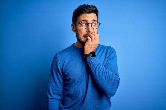 Young handsome man with beard wearing casual sweater and glasses over blue background looking stressed and nervous with hands on mouth biting nails. Anxiety problem.