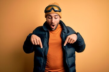 Young handsome skier man with beard wearing snow sportswear and ski goggles Pointing down with fingers showing advertisement, surprised face and open mouth