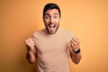 Young handsome man with beard wearing casual striped t-shirt over yellow background celebrating surprised and amazed for success with arms raised and open eyes. Winner concept.