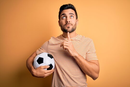 Handsome player man with beard playing soccer holding footballl ball over yellow background Thinking concentrated about doubt with finger on chin and looking up wondering