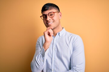 Young handsome hispanic business man wearing nerd glasses over yellow background looking confident at the camera with smile with crossed arms and hand raised on chin. Thinking positive.