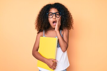 African american child with curly hair wearing glasses and holding book scared and amazed with open mouth for surprise, disbelief face