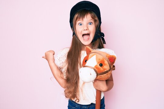 Little caucasian kid girl with long hair riding horse toy wearing vintage helmet pointing thumb up to the side smiling happy with open mouth