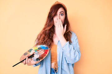 Young beautiful woman holding paintbrush and palette covering mouth with hand, shocked and afraid...