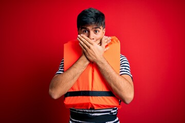 Young handsome man wearing orange safety life jacket over isolated red background shocked covering...