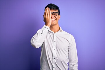 Fototapeta premium Young handsome business man wearing shirt and glasses over isolated purple background covering one eye with hand, confident smile on face and surprise emotion.