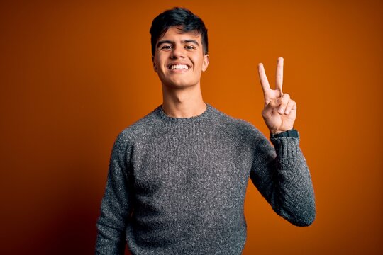 Young handsome man wearing casual sweater standing over isolated orange background smiling looking to the camera showing fingers doing victory sign. Number two.