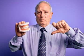 Grey haired senior man holding orthodontic prosthesis denture over purple background with angry...
