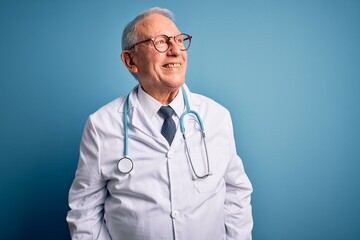 Senior grey haired doctor man wearing stethoscope and medical coat over blue background looking away to side with smile on face, natural expression. Laughing confident.