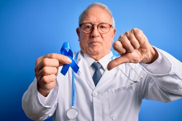 Grey haired senior doctor man holding colon cancer awareness blue ribbon over blue background with angry face, negative sign showing dislike with thumbs down, rejection concept