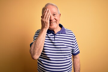 Grey haired senior man wearing casual navy striped t-shirt standing over yellow background covering...