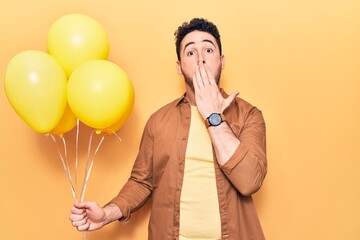 Young hispanic man holding balloons covering mouth with hand, shocked and afraid for mistake....