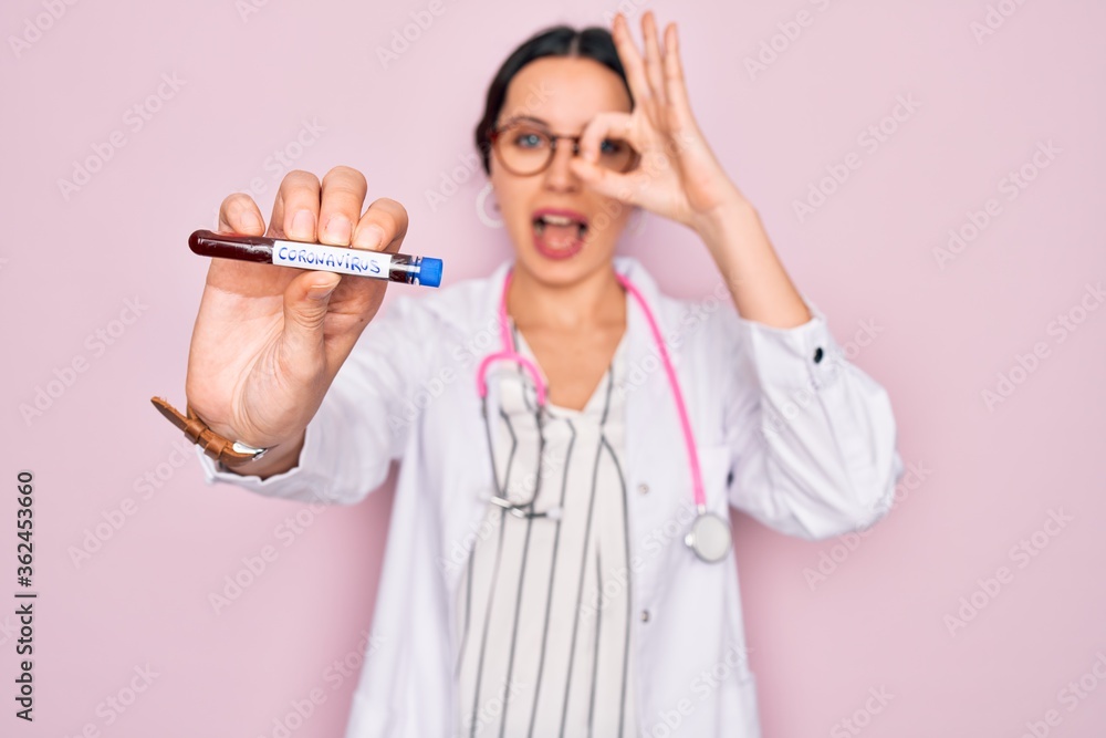 Wall mural Beautiful doctor woman with blue eyes wearing stethoscope holding coronavirus test tube with happy face smiling doing ok sign with hand on eye looking through fingers