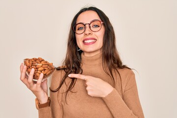 Beautiful woman with blue eyes holding bowl with healthy almonds over white background very happy pointing with hand and finger