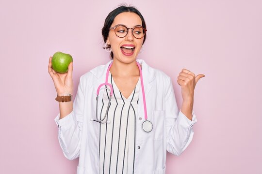 Young beautiful doctor woman with blue eyes wearing stethoscope holding green apple fruit pointing and showing with thumb up to the side with happy face smiling