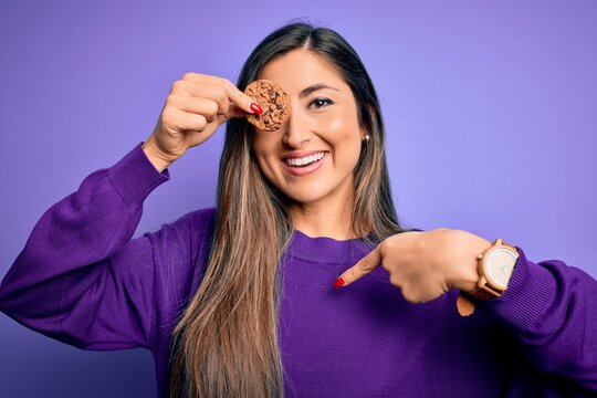 Young beautiful brunette woman holding healthy chocolate cookie over eye with surprise face pointing finger to himself