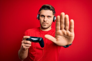 Young gamer man with blue eyes playing video games using gamepad joystick with open hand doing stop sign with serious and confident expression, defense gesture © Krakenimages.com