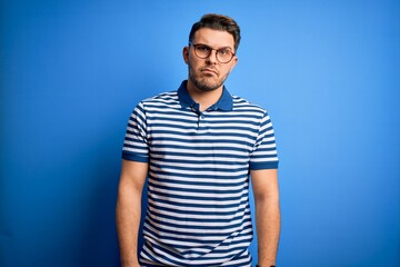 Young man with blue eyes wearing glasses and casual striped t-shirt over blue background skeptic and nervous, frowning upset because of problem. Negative person.