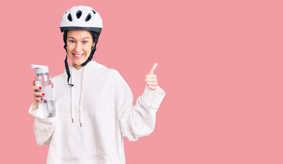 Beautiful brunette young woman wearing bike helmet and holding water bottle smiling happy and positive, thumb up doing excellent and approval sign