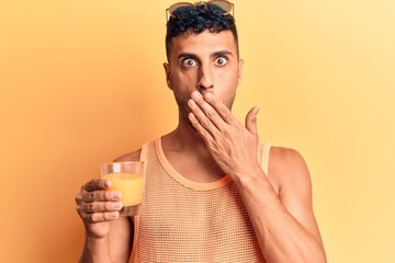 Young hispanic man drinking glass of orange juice covering mouth with hand, shocked and afraid for...