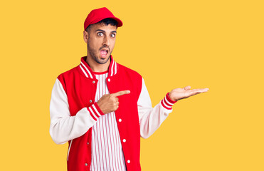 Young hispanic man wearing baseball uniform amazed and smiling to the camera while presenting with hand and pointing with finger.